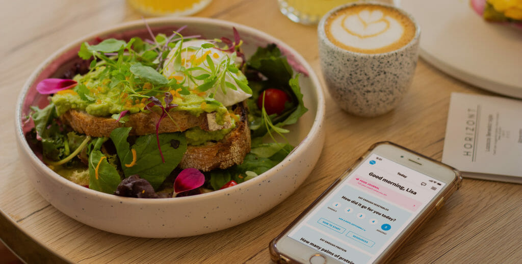 Healthy and colorful salad on a table next to a cafe latte and a smart phone featuring a nutrition app