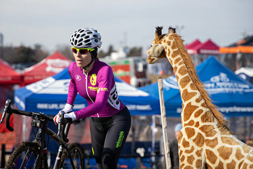 Theia Friestedt at cyclocross race pushing her bike up hill with stuffed giraffe spectating